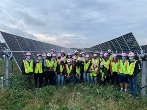 25 or so students in yellow jackets and hardhats in between two large solar panels