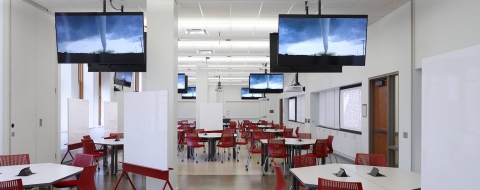 A large room with tables and chairs for collaborating with many TV screens and white boards on wheels.