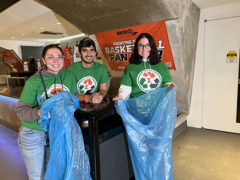 A group of 3 volunteers holding blue recycling bags near a trash bin.